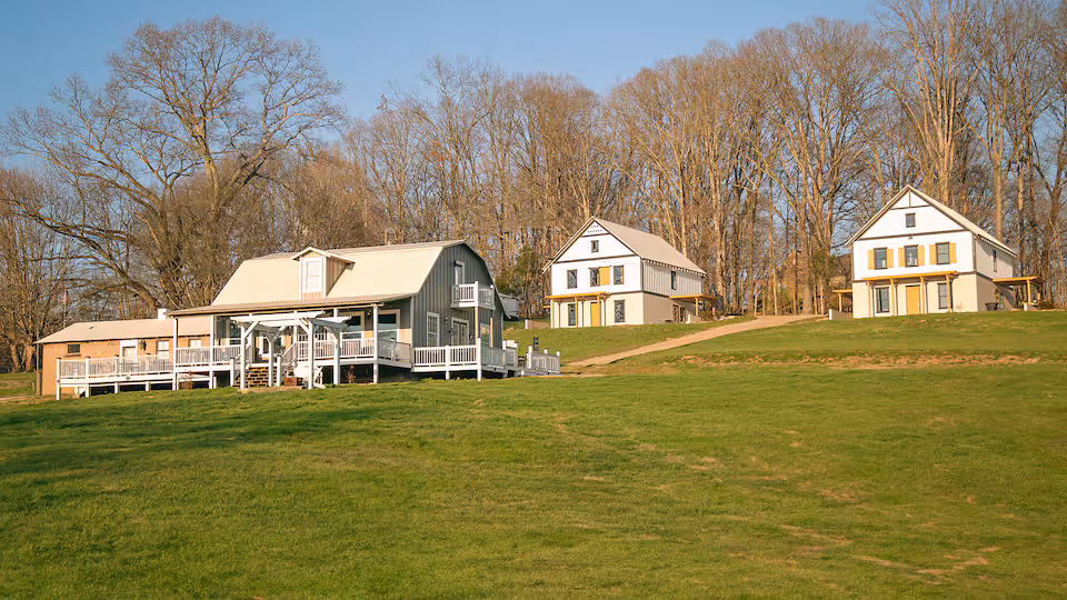 Three historic buildings sit on a grassy hillside surrounded by bare trees on a clear day.